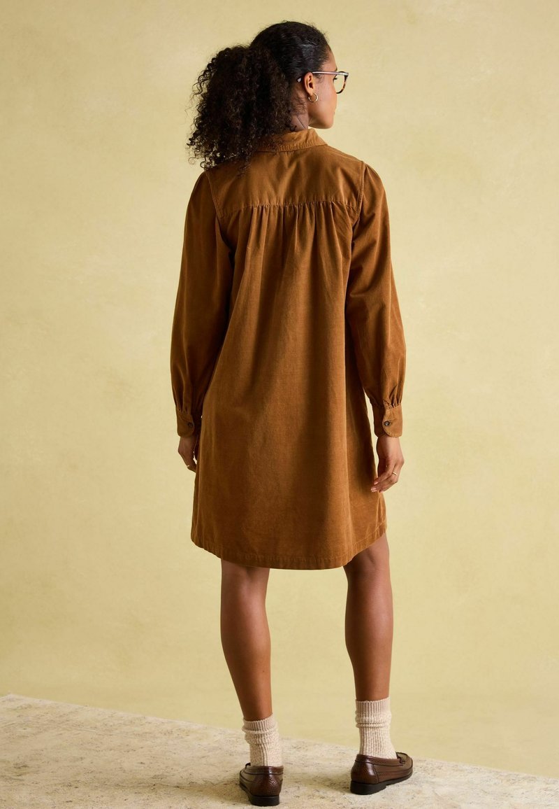 Braunes Cordhemdkleid mit langen Ärmeln, gerafftem Rückendetail und Knopfmanschetten, kombiniert mit hellen Stricksocken und dunklen Schuhen.