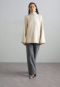 Μπεζ υπερμεγέθη πουλόβερ με ψηλό λαιμό και φαρδιές μανσέτες, συνδυασμένο με γκρι χαλαρές τζιν και μοτίβο ψηλοτάκουνα παπούτσια.