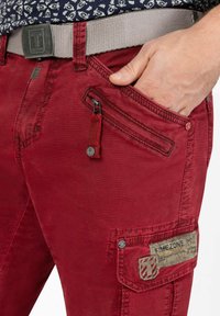 Red cotton pants with a rugged texture, featuring multiple pockets, a zipper, and a woven gray belt with a rectangular buckle.