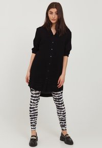 Schwarzes Hemdkleid mit Kragen und hochgekrempelten Ärmeln, kombiniert mit leggings in schwarz-weißer Musterung und klobigen schwarzen Schuhen.