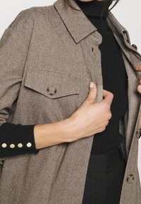 Brun herringbone-kappa med krage, framfickor och guldknappar på svarta ribbade ärmar. Nära bild av den texturerade tygen.