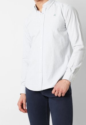 Mężczyzna ubrany w białą, drobno prążkowaną, zapinaną na guziki koszulę i ciemnoniebieskie spodnie, ręka spoczywająca blisko kieszeni, widoczny tylko tułów.