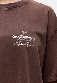 Brązowy, oversize t-shirt z graficznym motywem motyla oraz białym napisem "Goodfornothing". Miękka tekstura z delikatnymi różnicami kolorystycznymi.