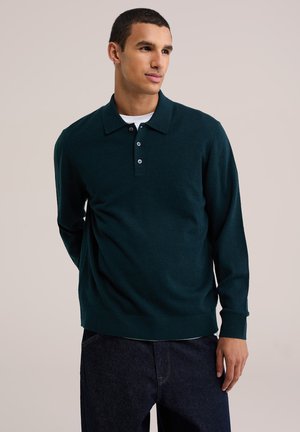 Jeune homme portant un polo à manches longues vert foncé et un jean foncé, regardant sur le côté avec les mains dans le dos sur un fond uni.