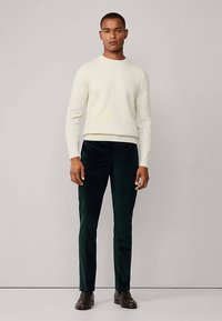 Pull en maille torsadée couleur crème clair, associé à un pantalon en velours côtelé vert foncé. Des chaussures marron complètent la tenue. Design simple et élégant.