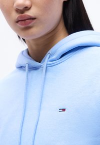 Κοντινό πλάνο ενός ατόμου που φοράει ανοιχτογαλάζιο φούτερ με κορδόνια και μικρό κόκκινο, άσπρο και ναυτικό λογότυπο στο στήθος.