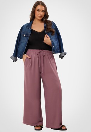 Donna che indossa pantaloni larghi color malva, top nero con rifiniture in pizzo, giacca di jeans appoggiata sulle spalle e sandali neri, in piedi con le mani nelle tasche.