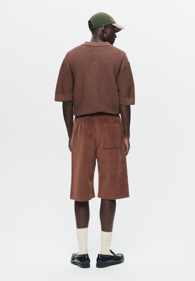 Chemise courte à manches courtes en tissu texturé marron et bermuda en velours côtelé assorti. Des chaussures foncées et des chaussettes crème complètent la tenue. Une casquette ajoute une couleur contrastante.