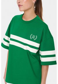 Maglietta verde a maniche corte in cotone, con strisce orizzontali bianche e un logo circolare con "SAINT TROPEZ 86." Texture liscia.