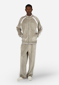 Beige kordbársony melegítő, fehér csíkokkal, cipzáras kabáttal és laza szabású nadrággal. Magas gallérral és oldalsó zsebekkel rendelkezik.