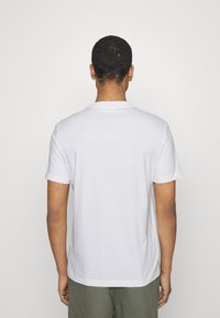 Camiseta blanca de algodón con cuello redondo, mangas cortas y una textura suave, mostrada desde la parte de atrás, combinada con pantalones verde oliva.
