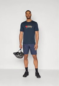 Tumši zila īsām piedurknēm T-krekls ar grafisku logotipu, kopā ar tumši ziliem šortiem. Figūra tur melnu velosipēda ķiveri. Valkātas melnas sporta kurpes.