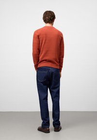 Uomo di spalle che indossa un maglione a maniche lunghe color ruggine, jeans blu scuro e scarpe marroni, in piedi contro uno sfondo semplice.