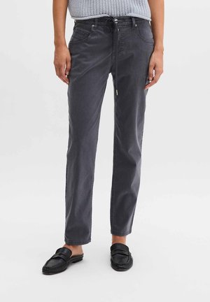 Person, der dunkelgraue Hose mit Kordelzug und schwarze Slip-on-Loafer trägt, steht vor einem einfarbigen hellen Hintergrund.