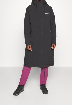 Personne portant un long manteau noir à capuche avec poches avant, pantalon magenta et chaussures de randonnée noires, debout devant un fond blanc uni.