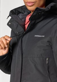 Czarna wodoodporna kurtka z zamkiem błyskawicznym z przodu, regulowanym kapturem i elastycznymi mankietami. Posiada zapinaną na suwak kieszeń i logo na piersi.