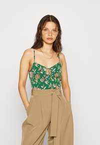 Camisola floral verde com alças finas, apresentando detalhes em rosa e dourado. Combinada com calças bejes de cintura alta, ajustadas, com um cinto amarrado.