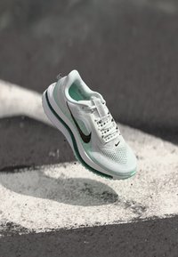 Chaussure de sport blanche et vert menthe avec une tige en maille, logo swoosh noir et semelle rembourrée, suspendue au-dessus d'une surface grise avec des marques blanches.