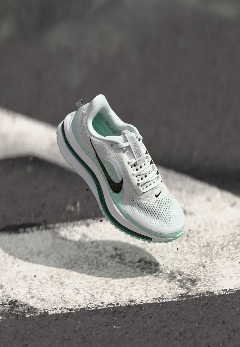 Chaussure de sport blanche et vert menthe avec une tige en maille, logo swoosh noir et semelle rembourrée, suspendue au-dessus d'une surface grise avec des marques blanches.