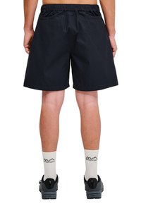 Shorts noirs avec une taille élastique, deux poches arrière et une coupe droite. Portés avec des chaussures grises et des chaussettes blanches comportant du texte.