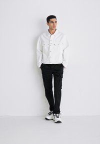 Weißes Denim-Jacket mit zwei Brusttaschen, getragen über schwarzen Cargo-Hosen und weißen Sneakers. Einfaches Design und taillierte Passform.
