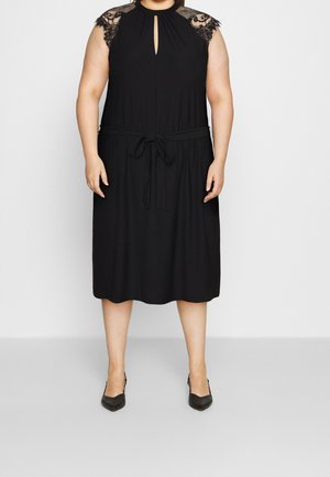 Femme portant une robe noire longueur genou avec des manches courtes en dentelle, un décolleté en trou de serrure à l’avant et une ceinture nouée à la taille, associée à des talons noirs.