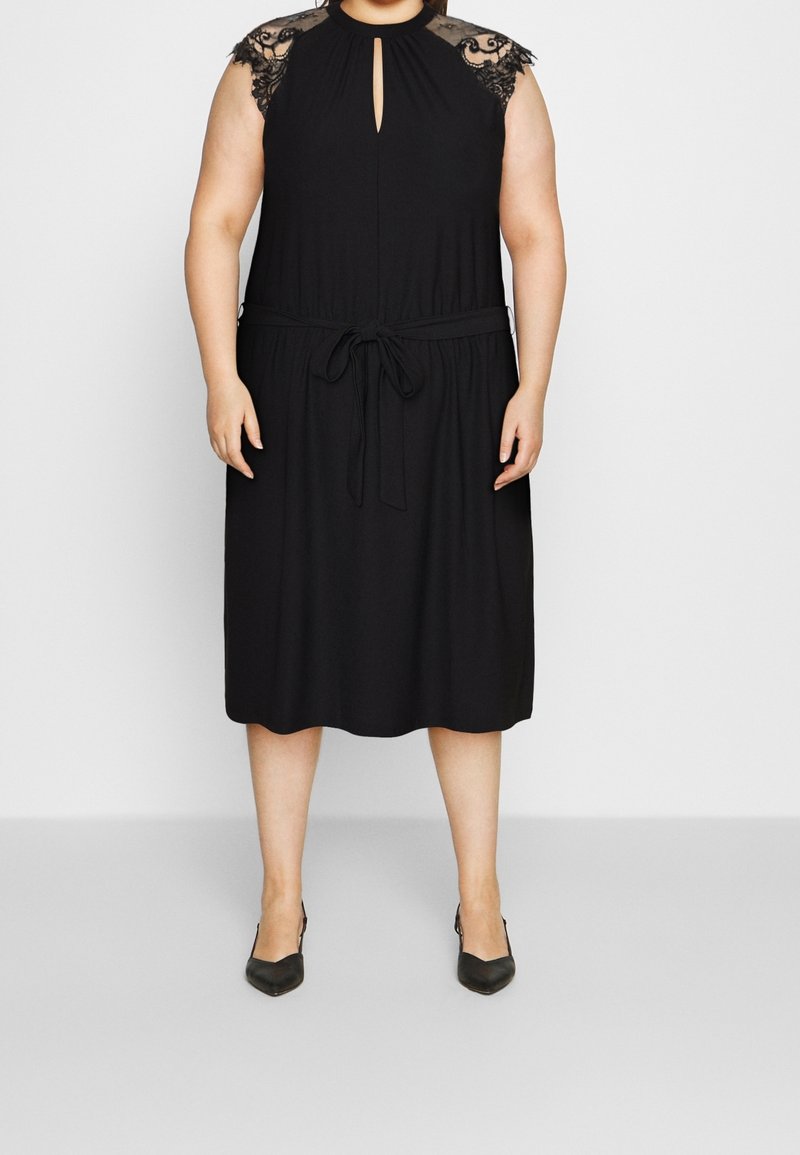 Femme portant une robe noire longueur genou avec des manches courtes en dentelle, un décolleté en trou de serrure à l’avant et une ceinture nouée à la taille, associée à des talons noirs.