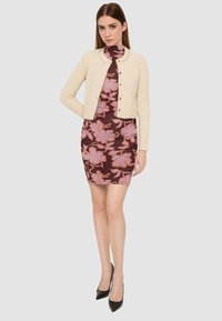 Cardigan corto beige con colletto rotondo e bottoni sopra un vestito attillato marrone a fiori con foglie rosa. Tacchi a punta neri completano il look.