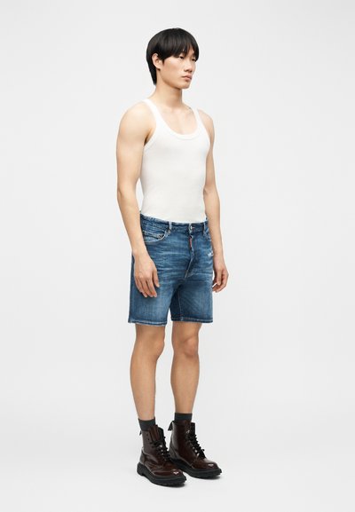 Jeune homme debout, portant un débardeur blanc sans manches, un short en jean bleu, des chaussettes noires et des bottes à lacets marron foncé sur un fond clair uni.