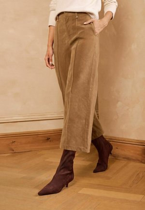 Personne portant un pantalon beige à jambes larges et des bottines à talons marron à bout pointu, debout sur un sol en bois contre un mur beige.
