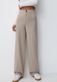 Beżowe spodnie szerokie, wykonane z gładkiego materiału, z zapięciem na guzik i dwiema przednimi kieszeniami. W zestawieniu z białymi trampkami.