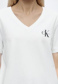 Γυναίκα που φοράει λευκό T-shirt με V-λαιμόκοψη και μικρό μαύρο λογότυπο Calvin Klein στο αριστερό στήθος.