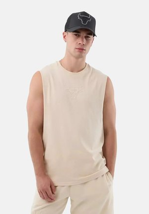 Jeune homme portant une chemise sans manches beige et un pantalon, casquette noire avec un logo de taureau, debout avec une main dans la poche devant un fond uni.