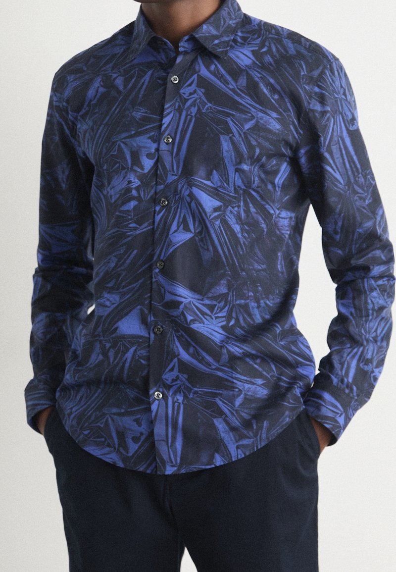 Homme portant une chemise à manches longues boutonnée avec un motif géométrique abstrait bleu brillant et un pantalon sombre, mains dans les poches.