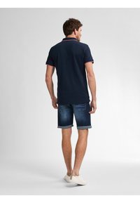 Námořnické polo tričko s červeným akcentem na límci, spárované s vykroucenými denimovými šortkami a béžovými slip-on botami, pohled zezadu.