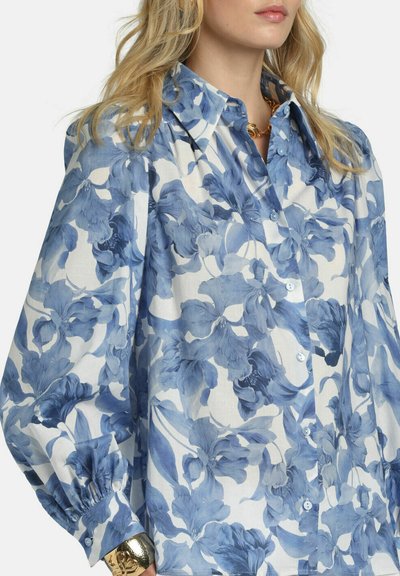 Woman wearing a white button-up blouse with blue floral print, puffed sleeves, and a gold bracelet and chain necklace.