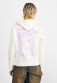 Sweat à capuche couleur crème avec un grand graphisme violet au dos, représentant des éléments de la nature et du texte. Poignets et taille côtelés.
