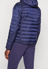 Navyblauwe gewatteerde jas met capuchon, met geweven secties en een elegante textuur. Logo op de schouder voegt detail toe; gecombineerd met bijpassende broek.