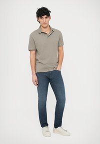 Svetlo siva polo majica z teksturo in ovratnikom, kombinirana s temno modrimi ozkimi kavbojkami in belimi supergami. Model stoji na enobarvnem ozadju.