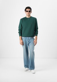 Πράσινη μπλούζα με στρογγυλή λαιμόκοψη, ανοιχτού μπλε παντελόνι wide-leg και λευκά αθλητικά παπούτσια. Τα αξεσουάρ περιλαμβάνουν στρογγυλά γυαλιά ηλίου. Απλό φόντο.