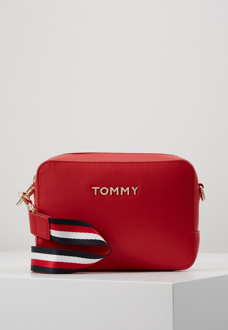 Czerwona prostokątna torba na ramię z złotym logo "TOMMY" i odczepianym paskiem w paski w kolorach czerwonym, białym i granatowym.