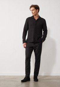 Cămașă button-up texturată neagră, pantaloni slim-fit negri și pantofi din suede negri. Modelul stă pe un fundal neutru, în fața unei fundaluri albe.