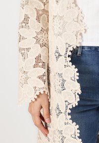 Beige lace cardigan with intricate floral patterns and scalloped edges. Worn over a white top and blue jeans. Light, sheer textile.