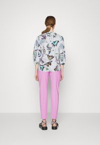 Blusa stampata con motivi di farfalle in tonalità di blu e verde, abbinata a pantaloni rosa. Il modello indossa sandali neri con cinturini.