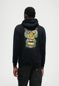 Hanorac negru cu un grafic detaliat pe spate, care prezintă o figură asemănătoare unei lei înconjurată de frunze verzi și flăcări.