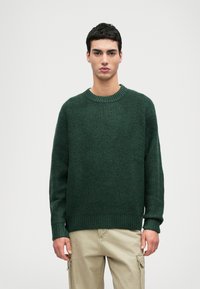 Mørkegrøn strikket sweater med en ribbet rund halsudskæring, lange ærmer og en afslappet pasform. Stof ser blødt ud med en tekstureret overflade.