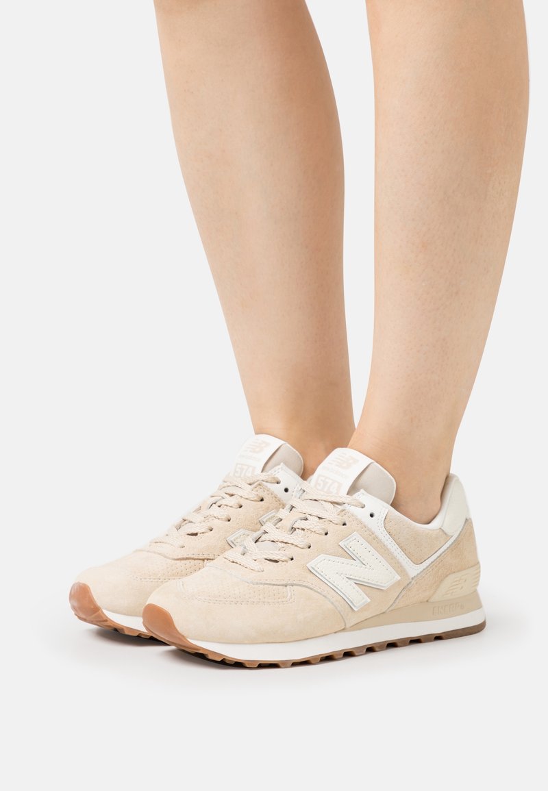 Pieds portant des baskets New Balance 574 beige avec des accents blancs et bruns sur un fond clair uni.