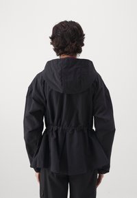 Chaqueta negra con capucha, cintura ajustada y detalle de espalda abullonada. Fabricada en tejido ligero, cuenta con puños elásticos y un corte relajado.