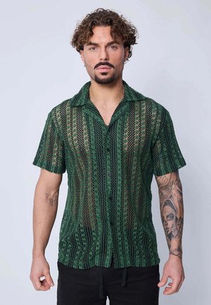 Homme aux cheveux bouclés et à la barbe portant une chemise verte semi-transparente à motifs à manches courtes et un pantalon noir, debout devant un fond uni.