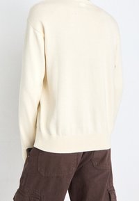 Ljust beige tröja med mjuk textur, rundad halsringning och ribbad fåll; kombinerad med mörkbruna cargo-byxor med sidofickor.